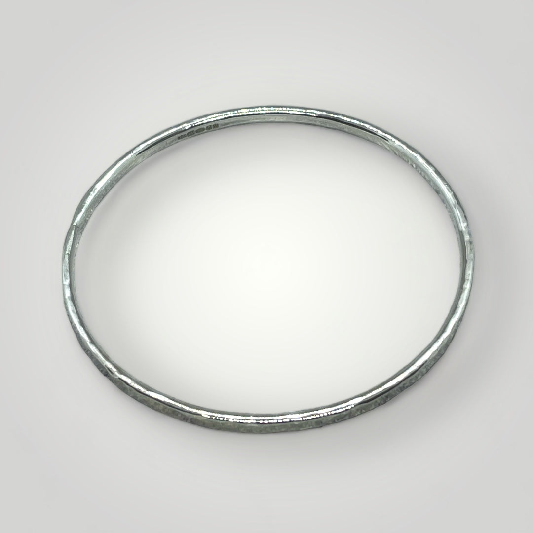 Handmade hammered sterling silver bangle on wrist, 3mm wide, hallmarked at Birmingham Assay Office, available in small, medium, and large sizes.