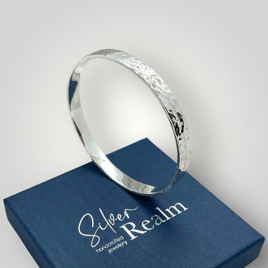 Angled view of a substantial handmade hammered sterling silver bangle showing solid silver thickness, on top of a Silver Realm branded gift box