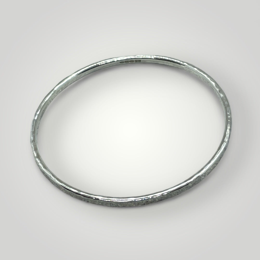 Handmade hammered sterling silver bangle on wrist, 3mm wide, hallmarked at Birmingham Assay Office, available in small, medium, and large sizes.