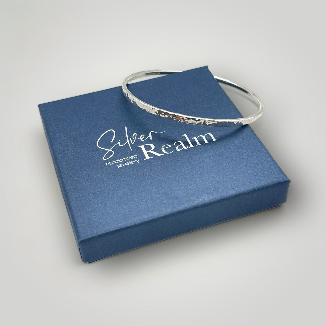 A silver bangle with a hammered finish, displayed on a blue box with 'Silver Realm' branding.