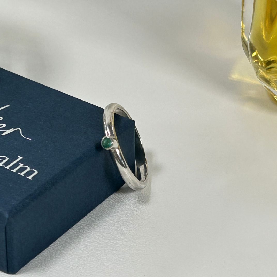 A sterling silver and Emerald stacking ring propped up against a blue, branded, gift box next to a bottle of perfume