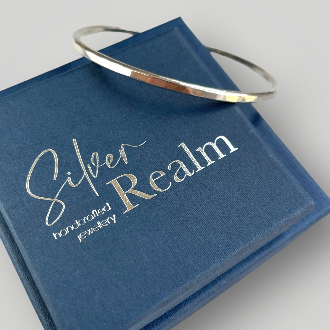 Handmade faceted sterling silver bangle displayed at an angle on a stack of Silver Realm Jewellery branded gift boxes