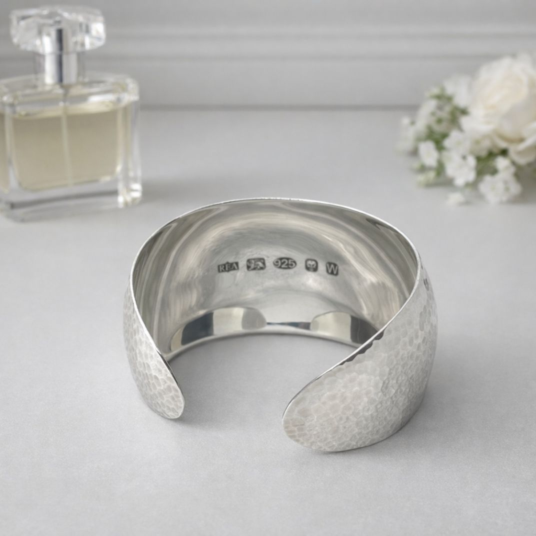 Handmade wide Argentium silver cuff bangle with hammered exterior, mirror-polished interior, and large feature hallmark on a light grey surface with a perfume bottle and flowers

