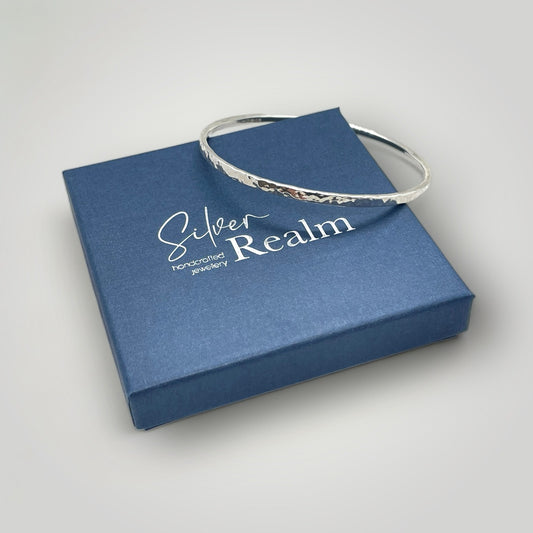Hammered sterling silver bangle resting on a Silver Realm branded gift box on a light grey background
