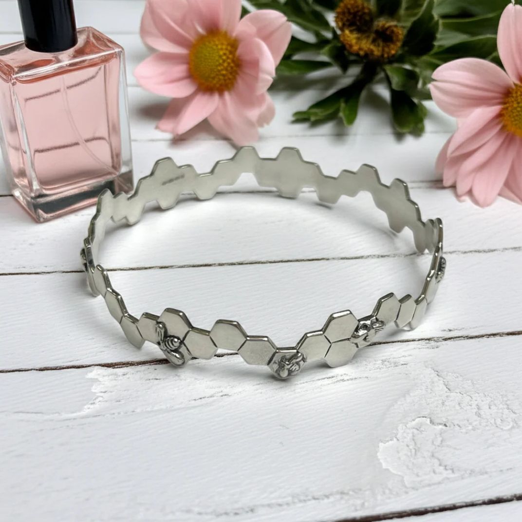 Sterling silver honeycomb and bee bangle on a white wooden surface with pink flowers and a perfume bottle in the background.