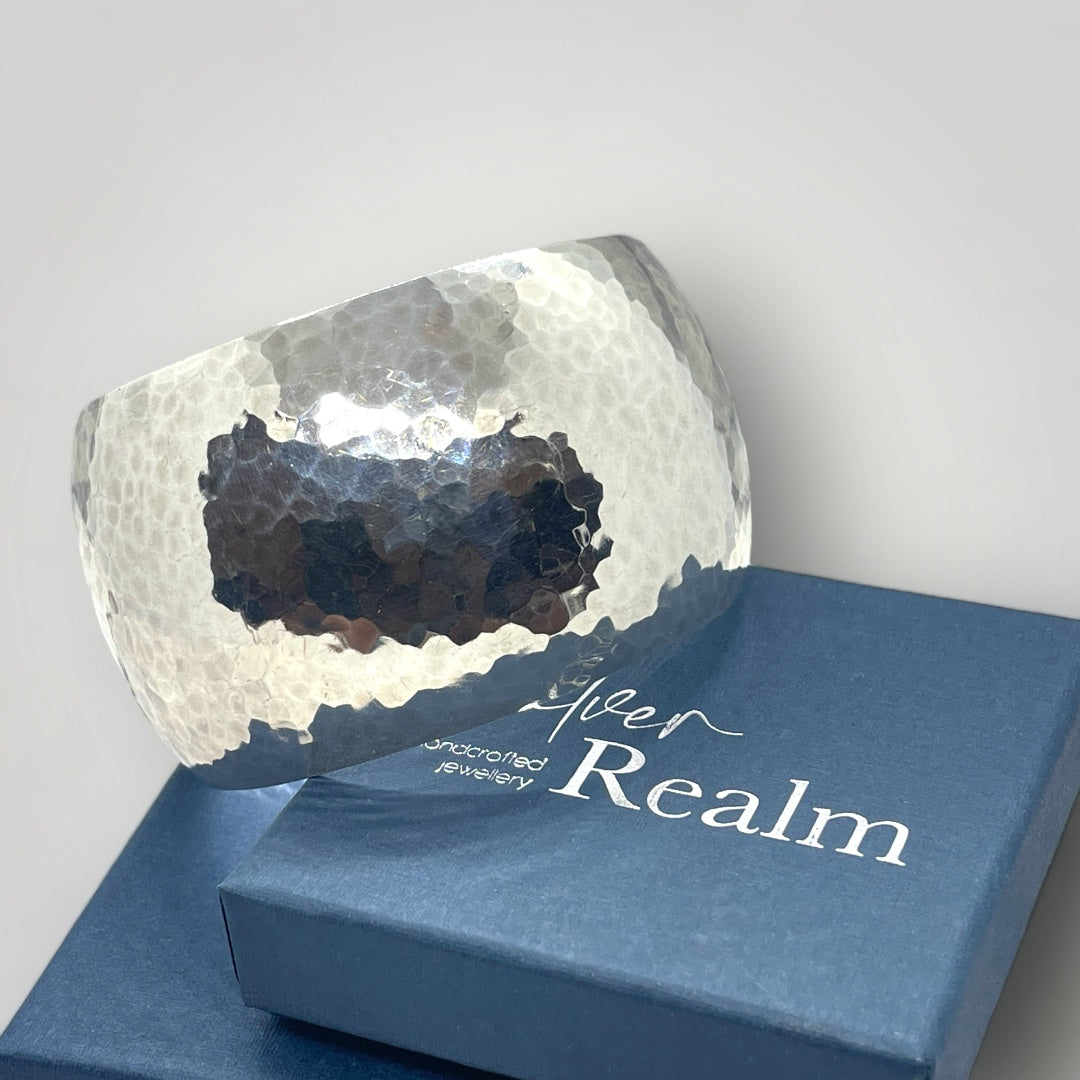 Handmade wide Argentium silver cuff bangle with hammered exterior, resting on a gift box with "Silver Realm" branding
