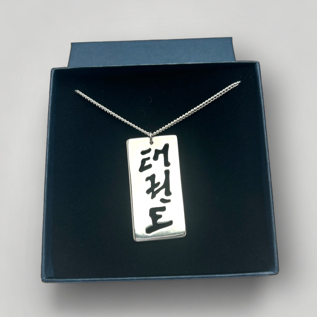 Sterling silver pendant with cut-out characters reading Taekwon-Do in Korean script in an open gift box on a gray background