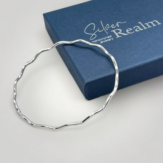 Wavy silver bangle on a white surface with a 'Silver Realm' branded box in the background.