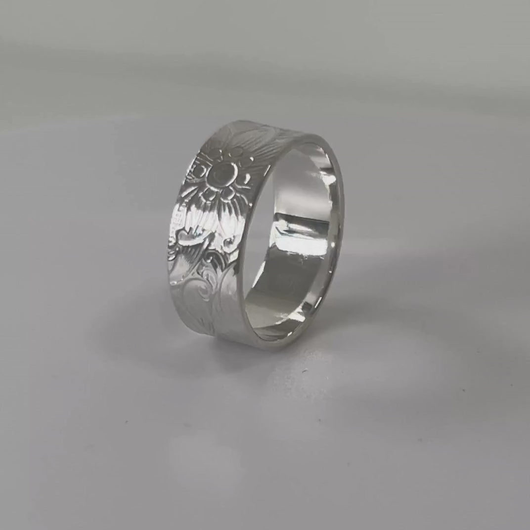 Textured sterling silver wide ring with floral and swirl detail rotating on a light grey background